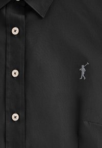 Maglietta nera con una texture liscia, caratterizzata da bottoni chiari e un piccolo logo grigio di una persona con un martello vicino alla tasca del petto.