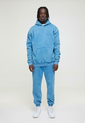 Blue hoodie and matching sweatpants set, made of soft fabric with a faded texture. Features a front pocket and elastic cuffs. Worn with white sneakers.