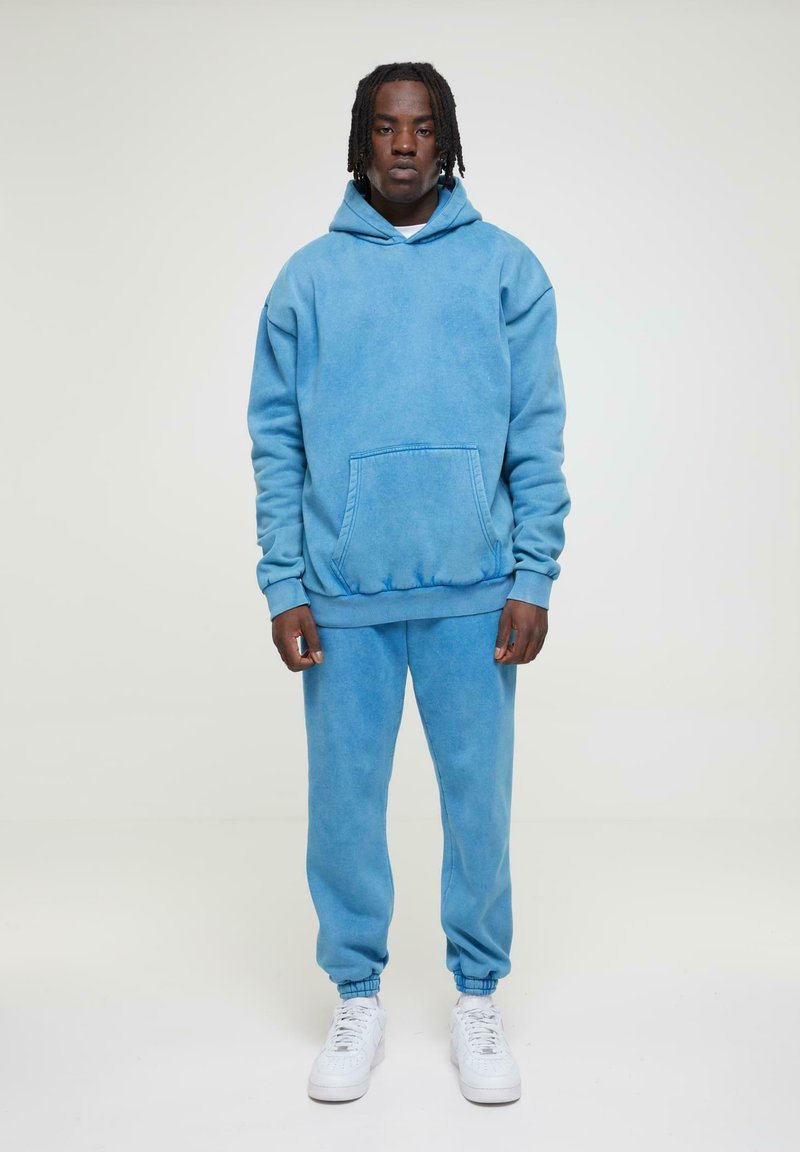 Ensemble hoodie bleu et pantalon de survêtement assorti, fabriqué en tissu doux avec une texture délavée. Dispose d'une poche frontale et de poignets élastiques. Porté avec des baskets blanches.