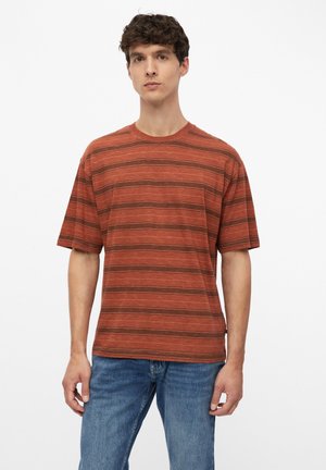 Jonge man die een roestoranje gestreept t-shirt en blauwe spijkerbroek draagt, staand tegen een effen witte achtergrond.
