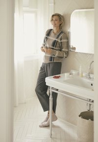 Pullover mit beigem und marineblauen Karomuster, graue Hose, helle Socken; weißes Waschbecken mit Hautpflegeprodukten, Fischgrätenfliesenboden.