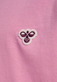 Różowy materiał z białym haftowanym logo przypominającym pszczołę, z ciemnopurpurowymi akcentami, umieszczonym na środku materiału.