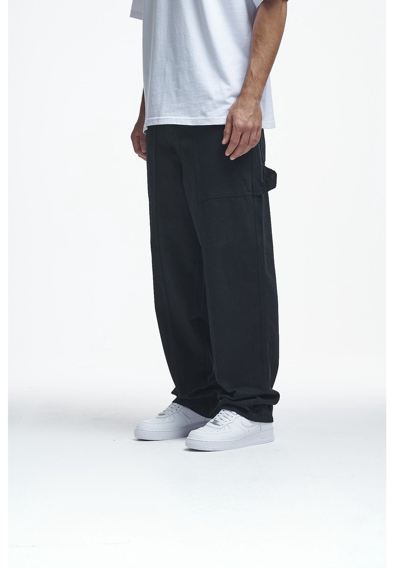 Homme portant un pantalon noir ample, des baskets blanches et un t-shirt blanc, debout devant un fond blanc uni.