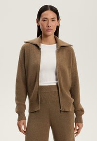 Cardigan tricotat maro cu guler înalt, fermoar complet, manșete cu textură și un model texturat, asortat cu un top alb ajustat.