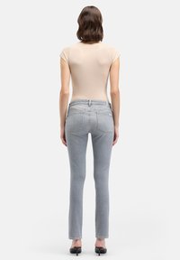 Kobieta stojąca tyłem, ubrana w beżową bluzkę z krótkim rękawem, jasnoniebieskie jeansy o dopasowanym kroju oraz czarne buty na obcasie na białym tle.