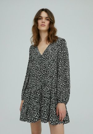 Robe noire avec motif floral blanc, manches longues, décolleté en V et ourlet à volants superposés. Fabriquée en tissu léger avec un design fluide.