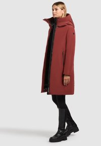Long manteau rouge avec un col haut, des zips et des poches sur les côtés. Porté sur un haut noir et associé à des bottes noires pour le contraste. Texture lisse.
