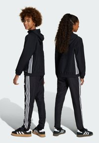 Svart idrottsoverall med en zip-up jacka med huva och matchande byxor. Båda har vita ränder längs sidorna. Sneakers ingår.