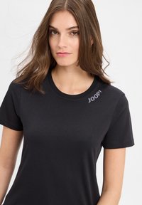 Zwarte katoenen t-shirt met korte mouwen, ronde halslijn, en een klein sprankelend logodetail op de linkerschouder. Gladde textuur.