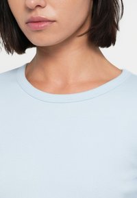 Majica svetlo modre barve iz rebrastega blaga s krožnim izrezom. Tekstura je gladka, majica pa ima prilegajoč se dizajn okoli vratu in ramen.