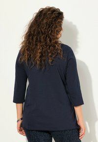 Marineblause Oberteil mit drei Viertelärmeln, lässiger Passform, glatter Textur und seitlichen Schlitz, kombiniert mit gemusterten dunklen Hosen.