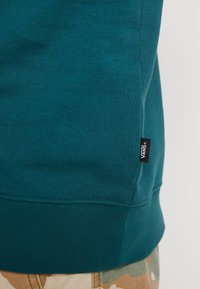 Sudadera en color verde azulado con un dobladillo acanalado, que presenta una pequeña etiqueta negra con el logo de Vans en el lateral. Material de mezcla de algodón con una textura suave.