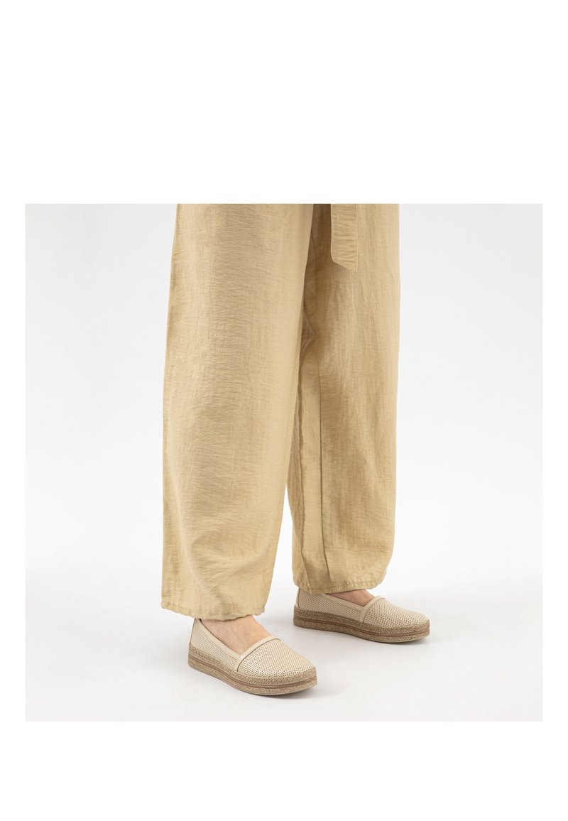 Beige gewebte Slip-On-Schuhe, getragen mit lockeren, hellbraunen, strukturierten Hosen mit weitem Bein auf weißem Hintergrund.