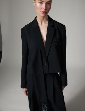 Kvinde iført en oversized sort blazer med en enkelt knap og matchende sorte bukser, stående mod en ensfarvet lys baggrund.