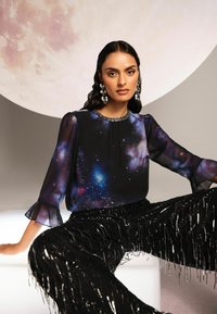 Fekete blúz galaxis mintával, áttetsző, trapézujjúval és gyöngyökkel díszített nyakkivágással. Fekete, rojtos flitteres nadrággal párosítva.