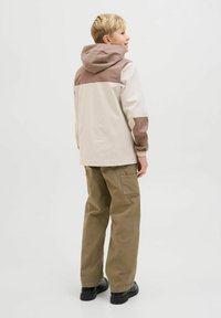 Giacca beige chiaro con accenti marroni sulle spalle e sulle maniche. Presenta un cappuccio, polsini elasticizzati ed è indossata con pantaloni cargo olivo e scarpe nere.