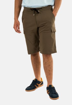 Pantalones cortos de cargo khaki con una cintura elástica, cordón y bolsillos laterales. Llevados con zapatillas oscuras con acentos naranjas.