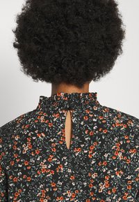 Svart blommig blus med en gatherad halsringning, liten nyckelhålsöppning bak och intrikata orange och vita blommönster genom hela.