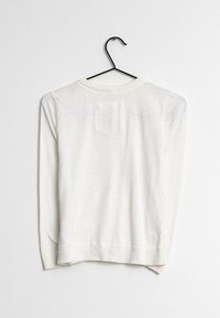 Maglione bianco a maniche lunghe appeso a un gancio nero, con scollatura rotonda, polsi e orlo a costine, e una piccola toppa quadrata sul retro.