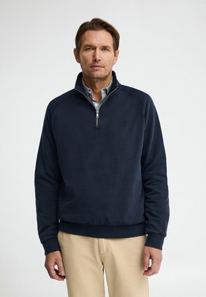 Uomo che indossa un pullover blu navy con mezza zip sopra una camicia azzurro chiaro e pantaloni beige, in piedi davanti a uno sfondo chiaro e semplice.