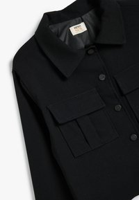 Camicia di cotone nera con colletto, dotata di due tasche sul petto, bottoni a pressione e una texture liscia. L'etichetta riporta "KIdston Kids."