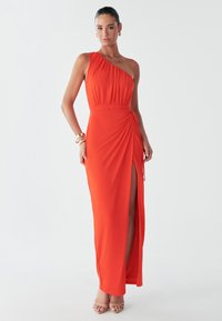 Rotes Maxikleid mit einer Schulter, seitlichem Schlitz, gerafftem Oberteil, Raffungen und glattem Stoffabschluss. Mit goldenen Schmuckaccessoires kombiniert.