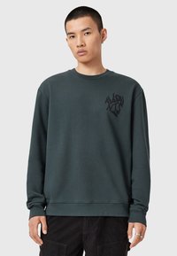 Tmavě zelená mikina s dlouhými rukávy, kulatým výstřihem a texturovaným černým logem na hrudi. Měkká tkanina s uvolněným střihem.