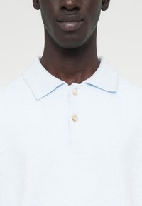 Ανοιχτό μπλε κοντομάνικο πόλο πουκάμισο από υφασμάτινη ύφανση, με κλασικό γιακά και τρία πέρλες κουμπιά στο λαιμό.