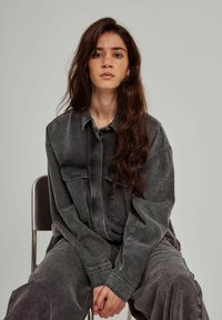 Grau denim Hemd mit langen Ärmeln, großen Taschen und Knopfverschluss; getragen mit passenden grauen Hosen. Schlichter Hintergrund, sitzende Person.