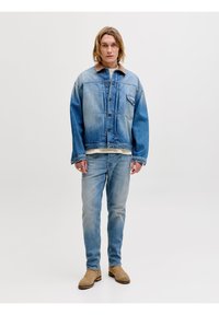 Νεαρός άνδρας που στέκεται φορώντας ξεθωριασμένο γαλάζιο τζιν μπουφάν, ανοιχτόχρωμο τζιν, μπεζ σουέτ μποτάκια και δαχτυλίδια στα δάχτυλα, μπροστά από απλό φόντο.