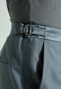 Sive krojene hlače izvode se od glatke, teksturirane tkanine s tamnim pojasom i metalnim detaljem kopče sa strane.