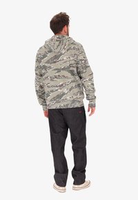 Uomo di spalle che indossa una felpa camouflage a strisce tigre verde e grigia, jeans scuri e sneakers bianche su uno sfondo bianco uniforme.