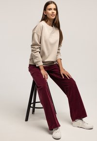 Beżowa bluza z wytłoczonym logo „LEE COOPER”; w połączeniu z bordowymi spodniami sztruksowymi; białe trampki; siedzi na czarnym stołku.