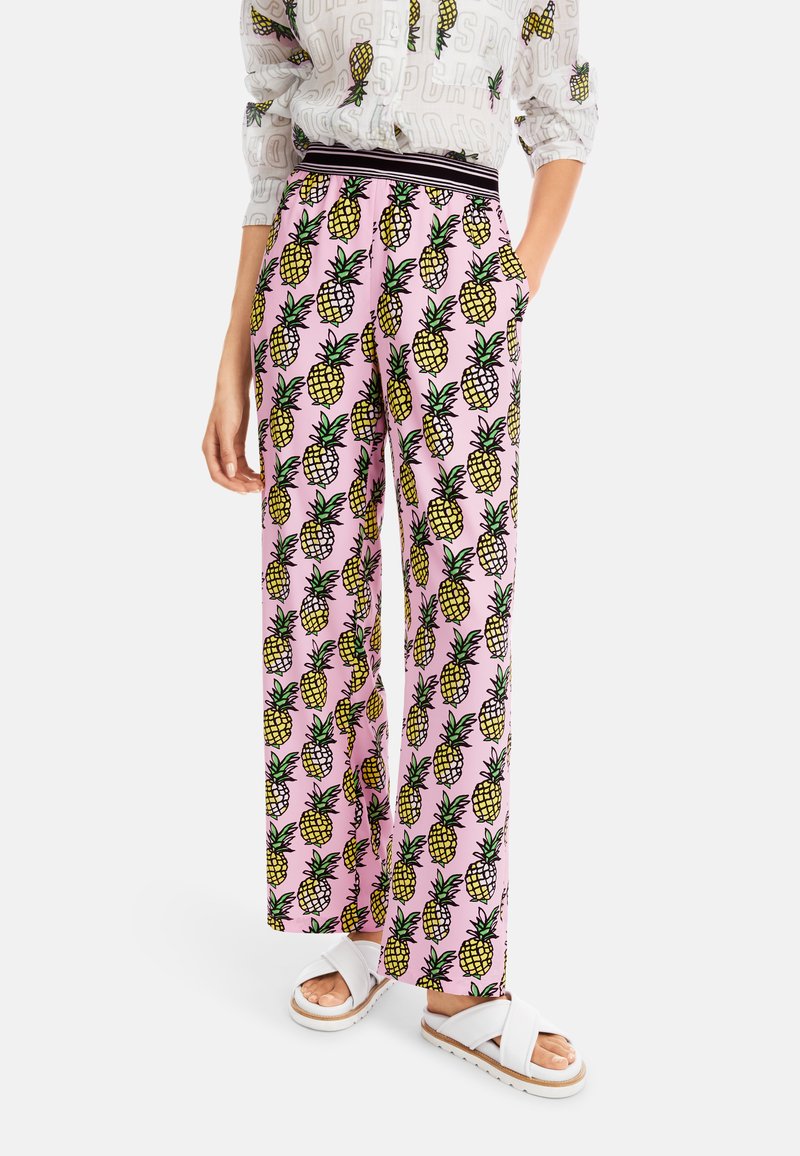 Rosa weite Hosen mit einem sich wiederholenden gelben Ananas-Muster, kombiniert mit weißen Sandalen. Das Material scheint leicht und lässig geschnitten zu sein.