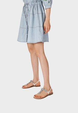 Donna che indossa un vestito azzurro chiaro al ginocchio e sandali bianchi piatti con strass decorativi, in piedi su sfondo chiaro.