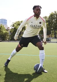 Fotbollskit med en krämfärgad långärmad topp, rosa detaljer och svarta shorts. Anmärkningsvärt logotyp: Qatar Airways. Spelare i aktion på en gräsmatta.