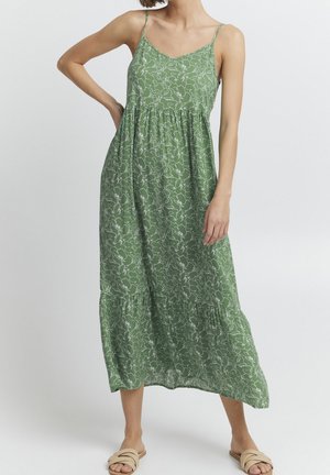 Kobieta w zielonej, kwiatowej sukience midi bez rękawów z cienkimi ramiączkami i beżowych sandałach na płaskim obcasie, pozuje z jedną ręką na biodrze.
