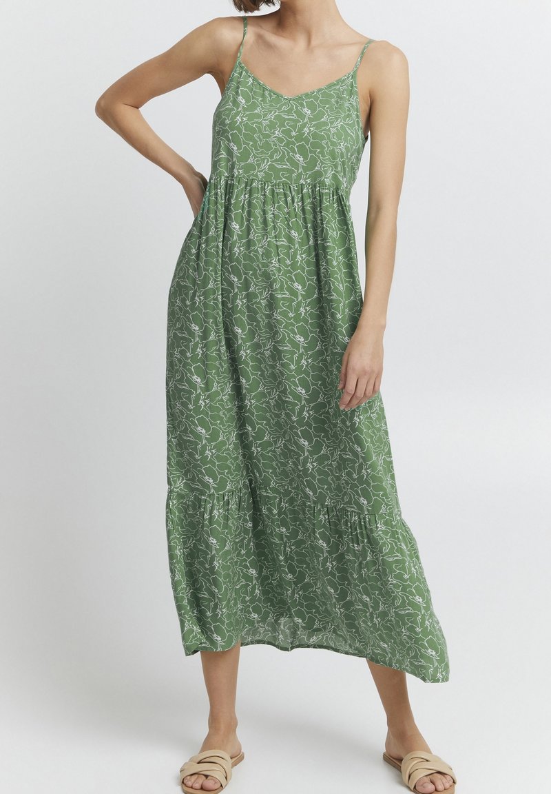 Femme portant une robe midi sans manches à fleurs verte avec de fines bretelles et des sandales beige à enfiler, posant avec une main sur la hanche.