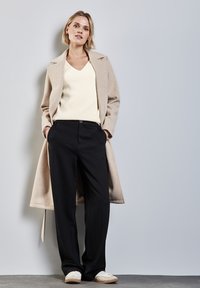 Cappotto lungo beige sopra un maglione a coste crema con scollo a V, abbinato a pantaloni neri e sneakers bianche. Il cappotto ha un colletto strutturato.