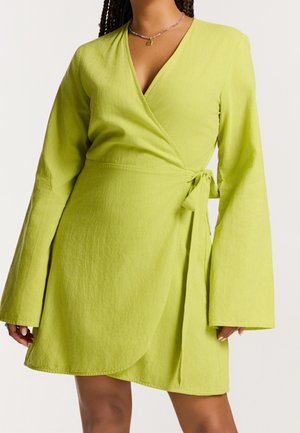 Femme portant une robe portefeuille vert citron avec de longues manches larges et une fermeture à nouer sur le côté, coupée au niveau de la moitié de la cuisse.