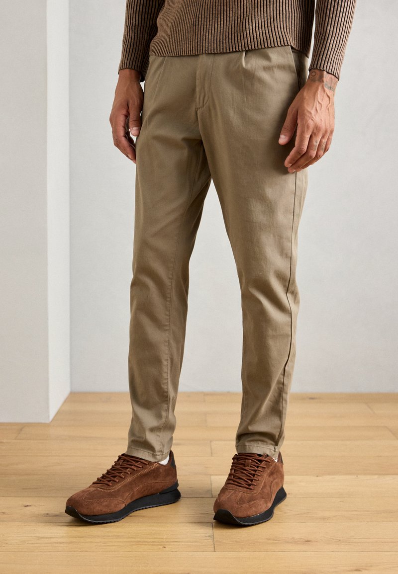 Man in beige chinos en bruine suède sneakers staand op een houten vloer tegen een effen muur.