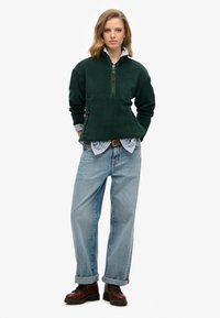 Donkergroen fleece pullover met een kwart-rits, voorzak en een kraag met een lichtblauw gestreept shirt, gecombineerd met baggy lichtblauwe jeans en bruine laarzen.