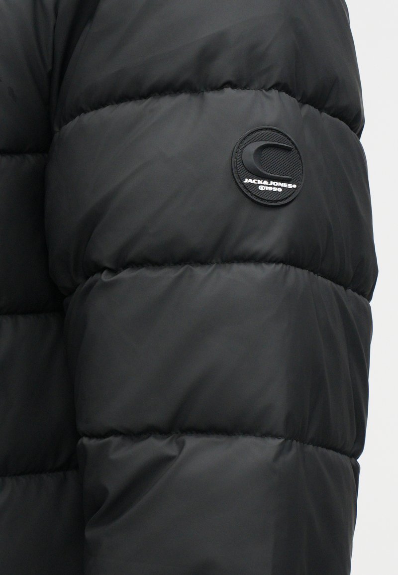 La veste doudoune noire présente un design matelassé avec une texture lisse et brillante. Elle comprend un patch logo circulaire avec le nom de la marque sur la manche.