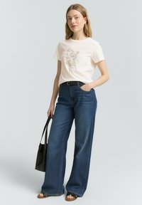 T-shirt en coton blanc avec un motif floral, associé à un jean en denim bleu à jambes larges. Une ceinture noire et un sac fourre-tout complètent le look. Des sandales plates sont portées.