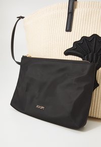 Trousse rectangulaire noire avec fermeture éclair et logo « JOOP ! » à l'avant, attachée à un sac fourre-tout beige tissé avec un écusson décoratif noir et des poignées en cuir.