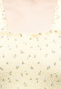 Κίτρινο φόρεμα από υφασμάτινη ύφανση με μικρό καφέ και λευκό λουλουδένιο σχέδιο και ραμμένη τετράγωνη λαιμόκοψη με βολάν, σε πρόσωπο.