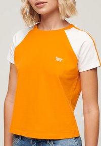 Camiseta de algodón naranja con mangas raglán blancas, cuello redondo y pequeño logo de Superdry en el pecho. Corte casual con una textura suave.