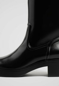 Stivale alla caviglia nero con finitura lucida, caratterizzato da un tacco a blocco e pannelli laterali elastici. Texture liscia e silhouette elegante.