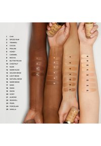 Drie armen met verschillende huidtinten tonen 25 swatches van foundationkleuren, elk gelabeld met een nummer en naam, terwijl ze foundationflessen vasthouden.