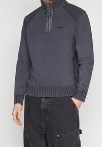 Polaire gris foncé avec col zippé, encolure et ourlet côtelés, portant un logo sur la poitrine. Coupe décontractée avec manches raglan.